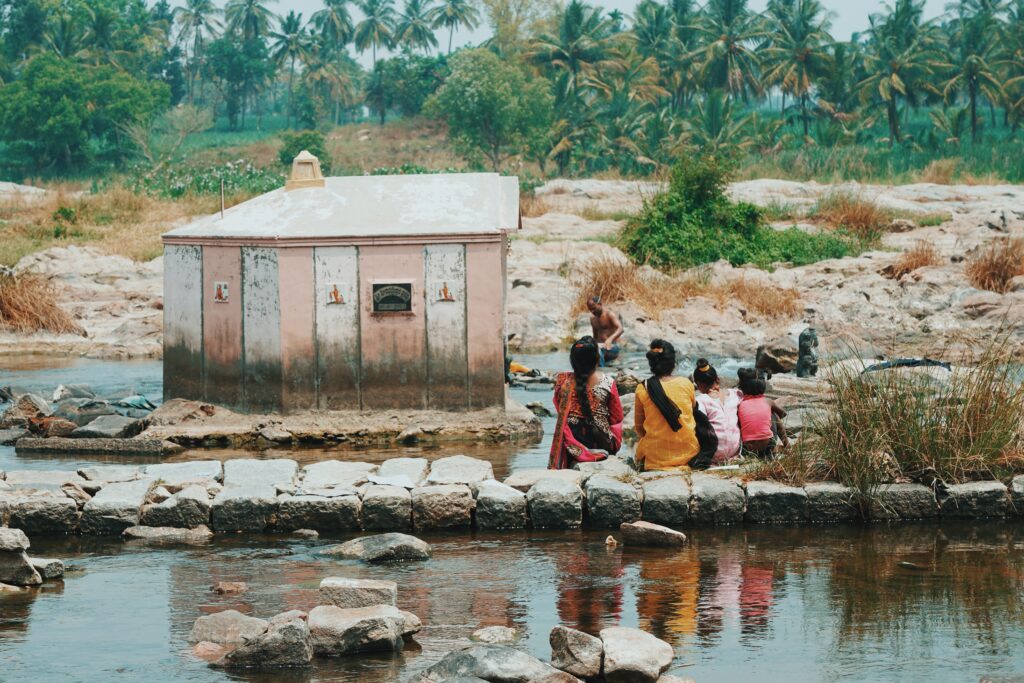 Row of women sitting next to a river facing a small structure.